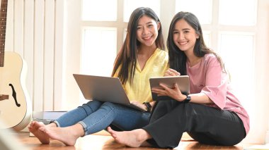 İki güzel kadının tablet ve dizüstü bilgisayarla rahatlarken rahat bir müzik stüdyosunun önünde yan yana oturduğu portre..