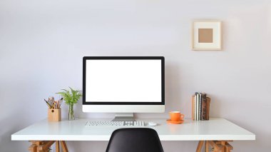 Kablosuz fare, klavye, kalem tutacağı, kitap yığını, kahve fincanı ve saksı bitkisini arka plan olarak beyaz duvarın üzerine koyarak çalışma masasına beyaz boş ekran koyan bilgisayar monitörü.