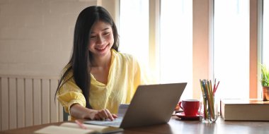 Güzel bir kadının, rahat oturma odasının pencereleri üzerinde ahşap masada otururken bilgisayar laptopu kullanmasının fotoğrafı. Öğrenim ve çevrimiçi öğrenim kavramı.