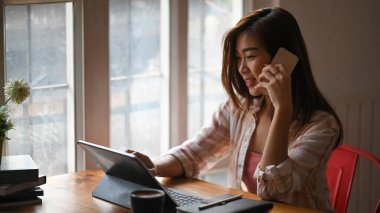 Güzel bir kadın cep telefonuyla konuşurken bilgisayar tableti kullanıyor ve ahşap masada oturuyor rahat oturma odasında arka plan olarak çalışıyor..