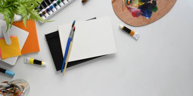 Ressam çalışma alanının en üstteki görüntüsü. Kırtasiye, renk paleti, boya fırçası, not defteri, renkli tüp ve saksı bitkisi bembeyaz çalışma masasında yatıyordu. Dağınık çalışma masası kavramı.