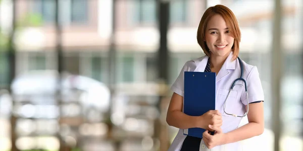 一个漂亮的女人 当医务工作者 面带微笑 手里拿着剪贴板 站在模糊的医院旁边做背景 — 图库照片