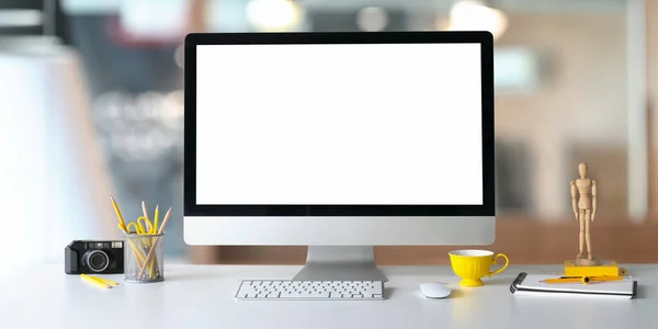 Photo of Computer monitor with white blank screen putting on white working desk and surrounded by camera, pencil holder, wireless keyboard, mouse, coffee cup, wooden puppet, note and dividers.