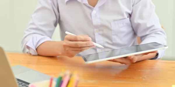 Cropped image of smart man in white shirt holding a computer tablet and stylus pen in his hands while sitting in front a computer laptop at the wooden working desk over living room as background.