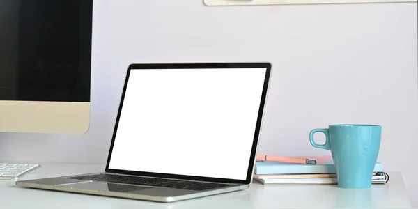 Photo of white blank screen computer laptop putting on white working desk surrounded by computer monitor, coffee cup and stack of books over white wall as background.