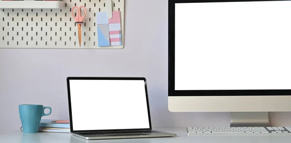 Photo of computer laptop with white blank screen putting on white working desk with coffee cup, stack of book, personal computer and keyboard over orderly living room wall as background.