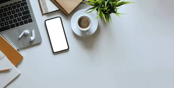 Top view image of smartphone with white blank screen putting on white working desk and surrounded by computer laptop, potted plant, notebook, diary, coffee cup, pencil and wireless earphone.