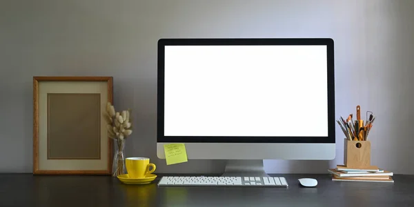 作業机の上に置かれ 絵のフレーム 鉛筆ホルダー 書籍のスタック ワイヤレスマウス キーボード コーヒーカップと花瓶の野生の草に囲まれたワークスペースの空白の画面コンピュータモニタの写真 — ストック写真