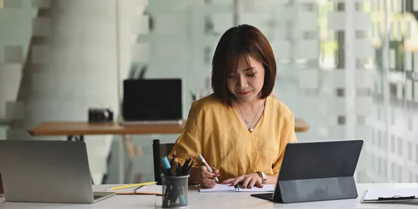 若い秘書女性書き込み紙の上に座っている間に彼女のコンピュータタブレットでキーボードケースとともにザ長い作業机の上に現代的なオフィスとして背景 — ストック写真