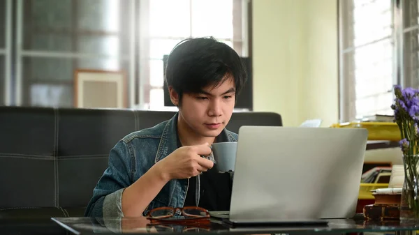 聪明的男人拿着热咖啡杯 用电脑笔记本电脑 坐在舒适客厅的皮沙发上作为背景的照片 — 图库照片