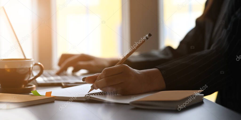 Cropped image of woman's hands writing on notebook and typing on laptop while sitting at the black working desk over modern sitting room as background.