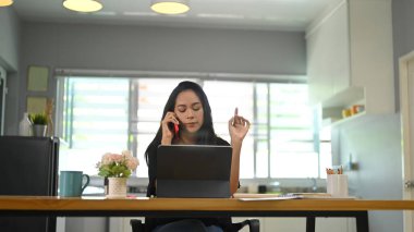 Genç bir kadın bilgisayar tableti kullanıyor ve ahşap masada otururken cep telefonuyla konuşuyor..