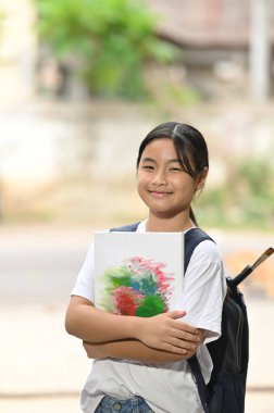 Bir kız öğrenci elinde boya malzemeleri ve okul çantasıyla okul otobüsünü bekliyor..
