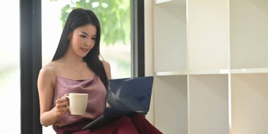 Güzel bir kadın kucağına koyduğu bilgisayara bakarken elinde kahve fincanı tutuyor..