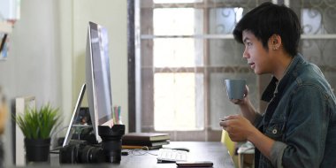 Akıllı bir adam çalışma masasında çalışırken kahve içer. Etrafı fotoğrafçı ekipmanlarıyla çevrilidir..