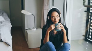 Kulaklıklı genç bir kadın yatak odasında otururken elinde kahve fincanı tutuyor..