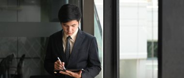 Bir iş adamı bilgisayar tableti kullanıyor ve modern ofisin arkasında duruyor..