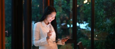 Bir iş kadını elinde bilgisayar tableti ve stilus kalemle ofis camının önünde dikiliyor..