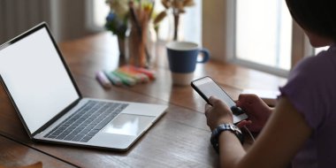 Güzel bir kadın ahşap çalışma masasında beyaz ekranlı bir bilgisayarın önünde otururken ve şarj olurken akıllı telefon kullanıyor..