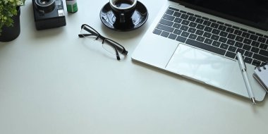 Laptop, kalem, bardak, kahve fincanı, not, retro kamera, film ve saksı bitkisi olan beyaz çalışma masası.