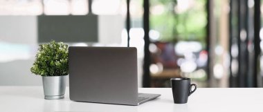 Çalışma alanı bilgisayarı, kahve fincanı ve saksı bitkisiyle çevrili beyaz bir masanın üzerine koyuyor..