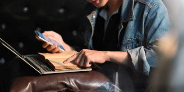 Akıllı bir adam elinde akıllı bir telefon tutarak kitap okuyor ve dizüstü bilgisayarın önünde oturuyor. Bu dizüstü bilgisayarla arka planda oturma odasında rahat bir kanepeye oturuyor..