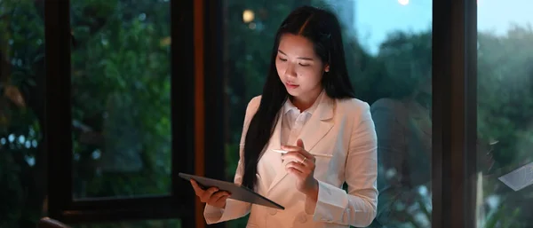 A businesswoman is holding a computer tablet and stylus pen while standing over the office glass wall.