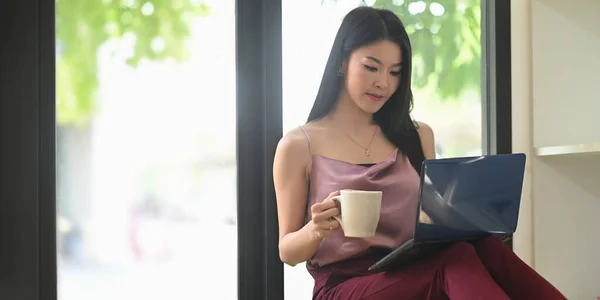 一位漂亮的女士一边拿着咖啡杯 一边看着放在膝盖上的笔记本电脑 — 图库照片