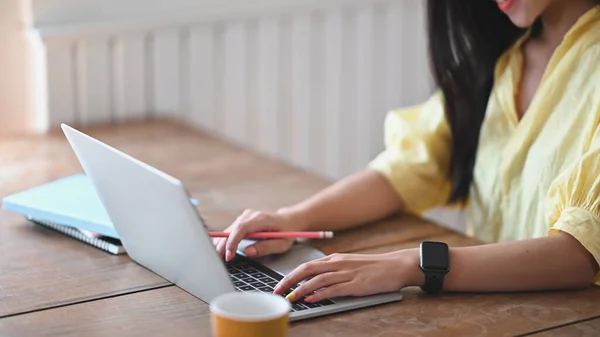 The young woman is taking notes while sitting in front of her computer laptop at the wooden working desk.