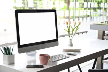 Beyaz masa üzerinde boş ekran bilgisayarı, kahve, kalem ve lale çiçeği olan yaratıcı çalışma alanı.
