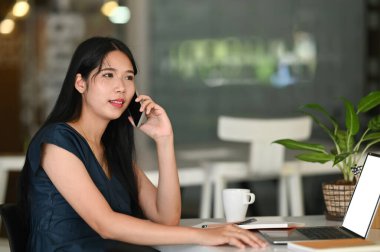 İşveren kadın akıllı telefonlu müşteriyle proje alımı siparişi hakkında konuşuyor. Çalışma alanındaki dizüstü bilgisayarını kullanarak..