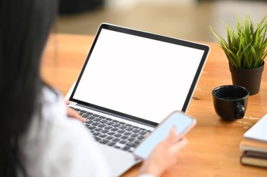 Ahşap masa üzerinde beyaz ekranlı dizüstü bilgisayar kullanırken çalışan kadınların akıllı telefonlarda daktilo yazma görüntülerini kapat.