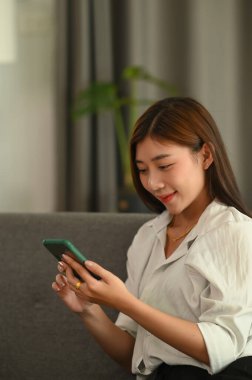 Evdeki koltukta otururken akıllı telefon kullanan genç bir kadının portresi..