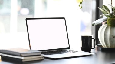 Minimal workplace with blank screen laptop, coffee cup and note books on white desk.