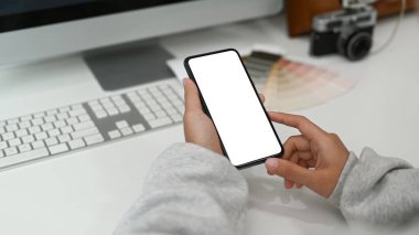 Cropped shot of holding blank screen smartphone on white desk with computer and office supplies at workspace.