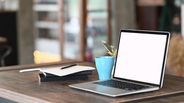 Computer laptop with white screen, coffee cup and notebook on working table in front of blur living room background.