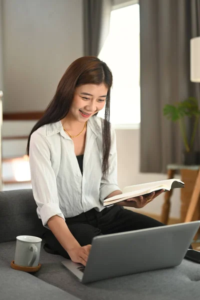 Szczęśliwa Kobieta Czytająca Książkę Podczas Korzystania Laptopa Kanapie Salonie — Zdjęcie stockowe
