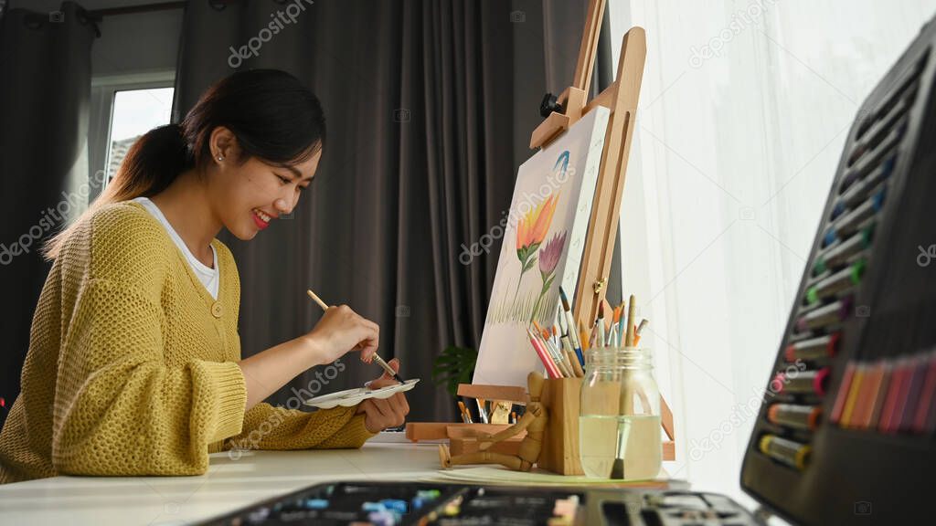 Portrait of happy young woman artist holding palette and paintbrush while working on painting in her workshop.