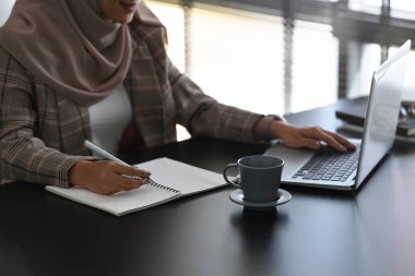 Başörtüsü takmış meşgul bir Müslüman kadın bilgisayar laptopuyla projesinde çalışıyor ve deftere notlar alıyor..