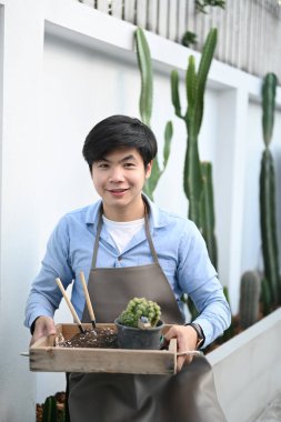 Evde kaktüsle tepsi tutarken kameraya gülümseyen genç bir adamın portresi..