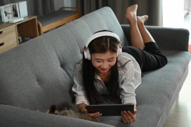 Kulaklıklı genç bir kız evde kanepede yatarken tablet kullanıyor ve müzik dinliyor..