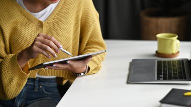Ofiste dizüstü bilgisayarda çalışırken dijital tablet ve stilus kalem kullanan genç bir kadının kırpılmış görüntüsü..