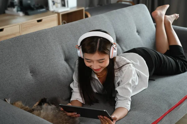 Młoda Kobieta Leży Kanapie Swoim Kotem Używając Tabletu Słuchawkami Salonie — Zdjęcie stockowe