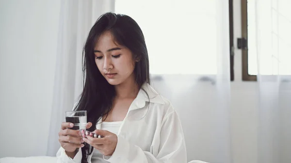 生病的女人坐在床上 手里拿着一杯水吃药 — 图库照片