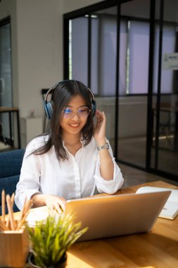 Kulaklıklı genç bayan tasarımcı gülümsüyor ve dizüstü bilgisayarla çalışıyor..