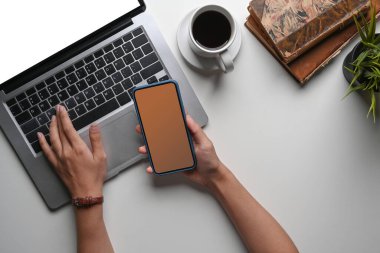 Dizüstü bilgisayarda çalışan ve masasında akıllı bir telefon tutan genç bir kadın..