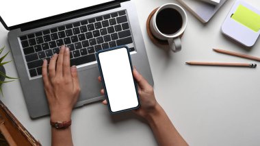 Akıllı telefon tutan ve ofis masasındaki dizüstü bilgisayarda çalışan kadının genel görünümü.