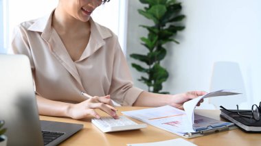 Bilgisayarla çalışan ve ofis masasında iş verilerini hesaplayan gülümseyen genç kadın muhasebeci..