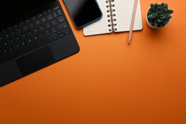 Masa bilgisayarı, akıllı telefon, not defteri ve turuncu arkaplanda kopyalama alanı olan tasarımcı çalışma yerinin üst görünümü.