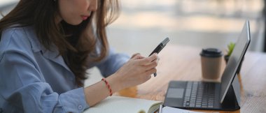 İş kadınının cep telefonu kullanırken ve ofiste bilgisayar tablosuyla çalışırken çekilmiş yatay fotoğrafı..